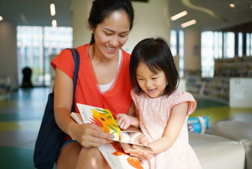 A mother and her daughter looking at a colourful picture book in the library.