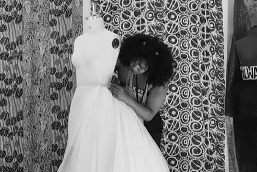A black and white photo of an Indigenous woman tailoring a white dress on a mannequin, in front of patterned curtains.