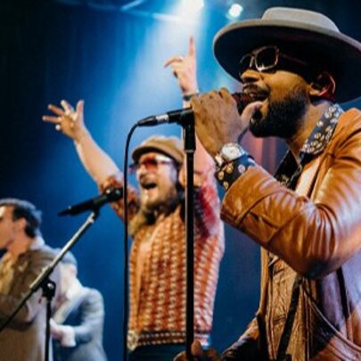 Singers performing lined up on stage in front of microphones, wearing sunglasses and hats.