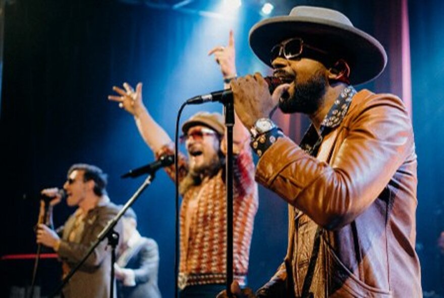 Singers performing lined up on stage in front of microphones, wearing sunglasses and hats.