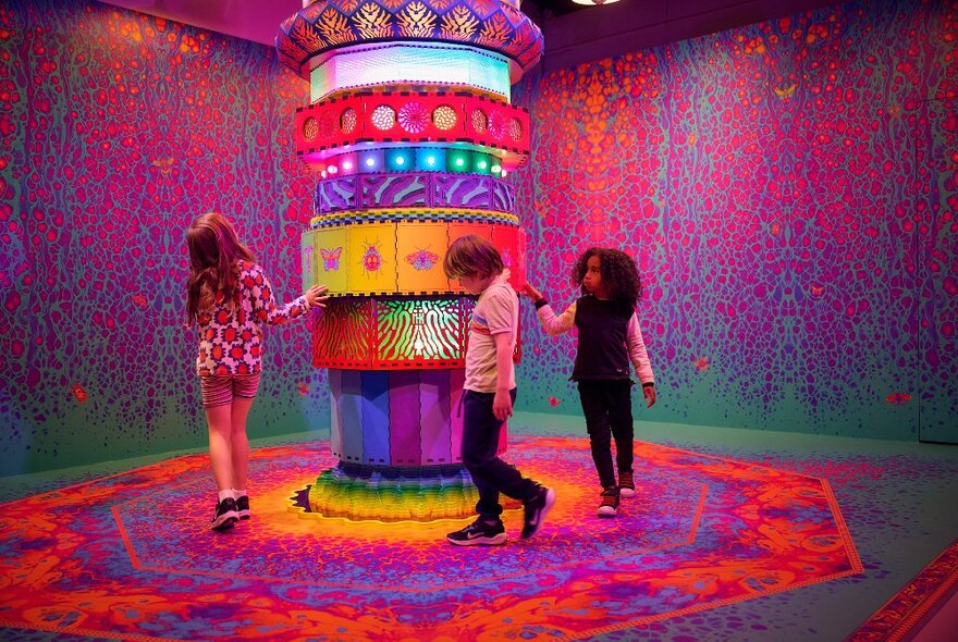 Three children circling a colourful pillar in an extremely colourful room in a gallery setting.