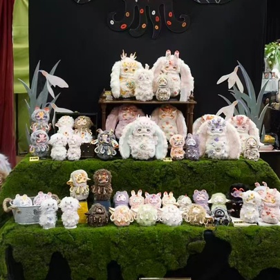 A collection of strange but cute statuettes of small animals displayed on a green moss-covered multi-tier stand at a market stall.
