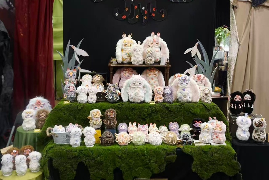 A collection of strange but cute statuettes of small animals displayed on a green moss-covered multi-tier stand at a market stall.