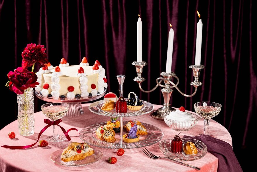 A selection of cakes and desserts arranged on a table covered in a pink tablecloth, with a candelabra and bunch of red flowers in a vase.
