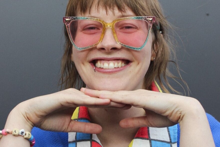A portrait of Alex Creece, smiling with their hands under their chin and wearing glasses.