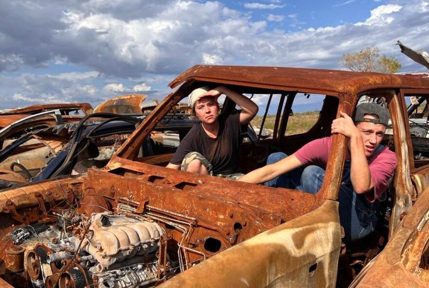 Two hound men in caps look confused in a rusted-out car in the desert. 