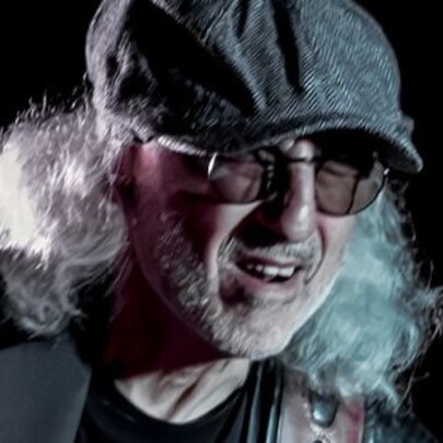 Blues musician Michael Charles, wearing glasses has has white shoulder length hair under a grey cap.
