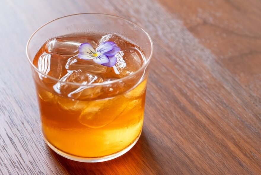 An amber drink in a tumbler with a mauve viola garnish. 