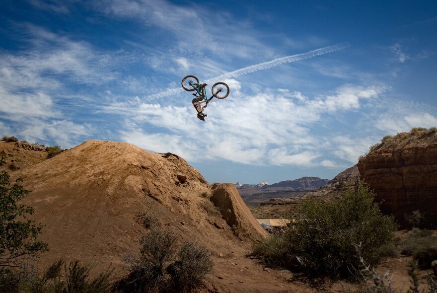 A stunt cyclist upside down on a bike in the air in a mountainous terrain. 