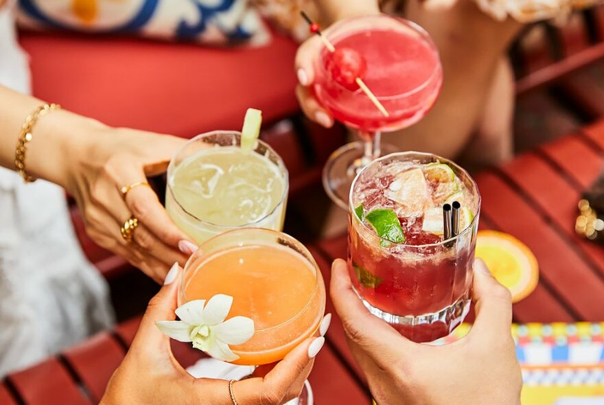 Four hands bringing four different coloured cocktails together in a cheers or clinking glasses motion.