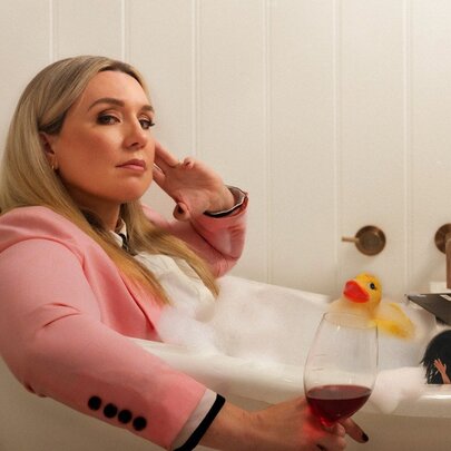 A woman wearing a pink blazer, holding a glass of red wine, in a bubble bath with a rubber duck.