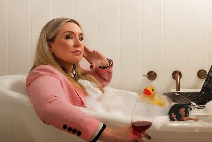 A woman wearing a pink blazer, holding a glass of red wine, in a bubble bath with a rubber duck.