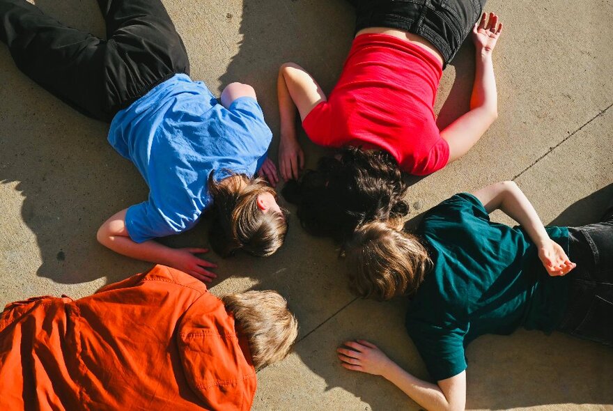 Four people lying face down on a concrete floor with heads together in a circle, each wearing a different brightly coloured top.