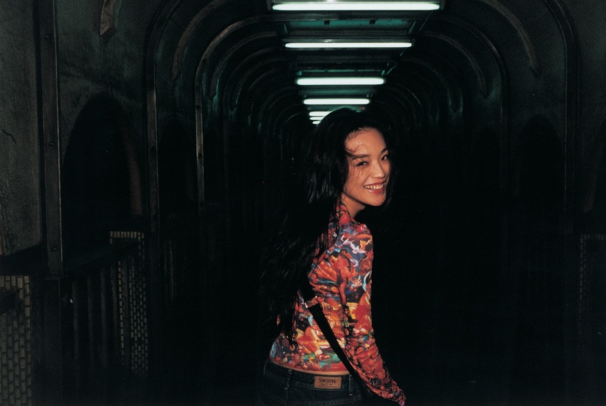 Movie still of a woman turning around, smiling, in a dark tunnel with overhead strip lighting.