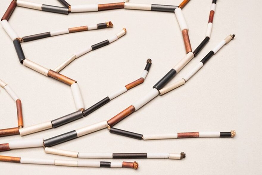 Short, cylindrical beads or components in white, copper, and blackened metal finishes, as part of a contemporary jewellery piece. 