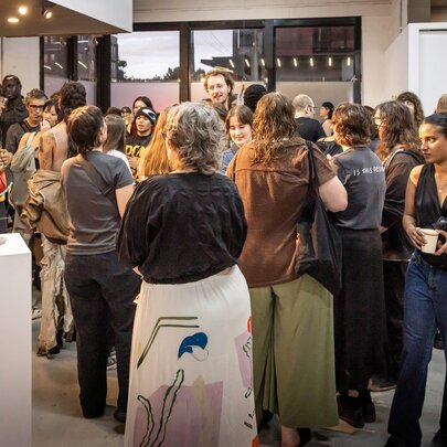 A crowded opening night at an art gallery or exhibition space with people standing around socialising and looking at artworks.