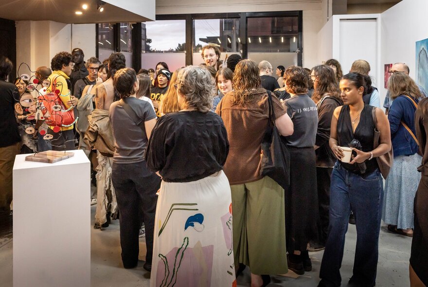 A crowded opening night at an art gallery or exhibition space with people standing around socialising and looking at artworks.