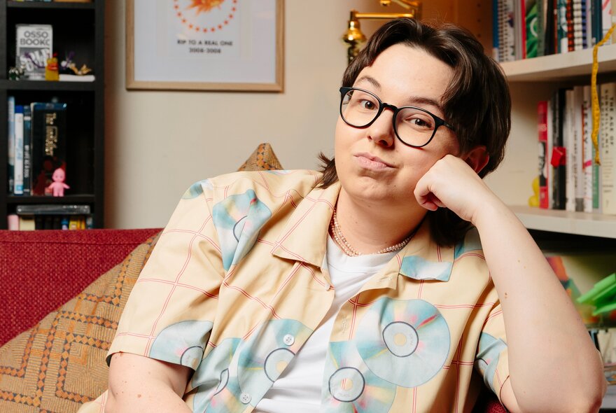 Comedian Maddy Weekes sitting in a room with bookshelves looking bemused leaning her head on her hand