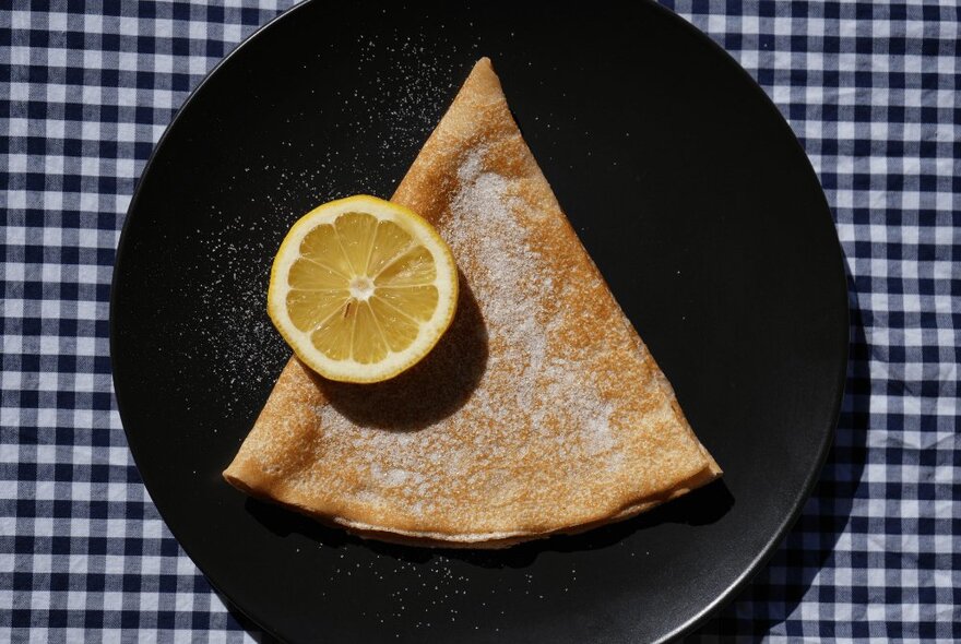 A classic triangular shaped French crepe with a slice of lemon on a black plate on a black gingham tablecloth.