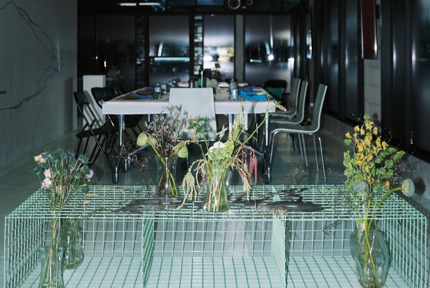 A modern interior design studio or a workshop, featuring a long table with chairs and multiple floral arrangements in vases.
