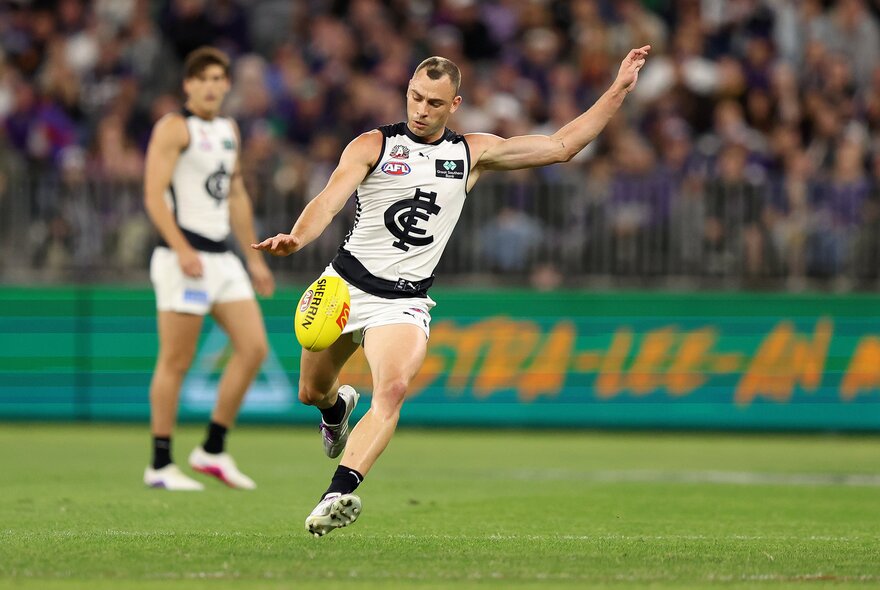 A Carlton AFL football player running and about to kick the footy during a match, with  blurred fans in the stands behind him.