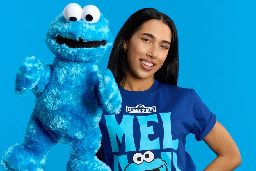 A person wearing a blue Sesame Street t-shirt and holding a large blue Cookie Monster soft toy.