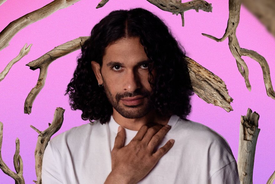 An actor with dark wavy hair wearing a white t-shirt and resting his hand on his upper chest, posed against a pink backdrop with broken tree branches arranged behind him.