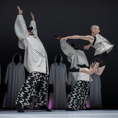 A dynamic modern dance performance on a dark stage, with cloaked figures in the background and three dancers centre stage, one leaping in the air.