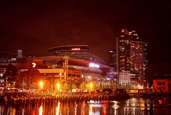 Docklands harbour lit up with flame jets during Firelight Festival, with Marvel Stadium visible.
