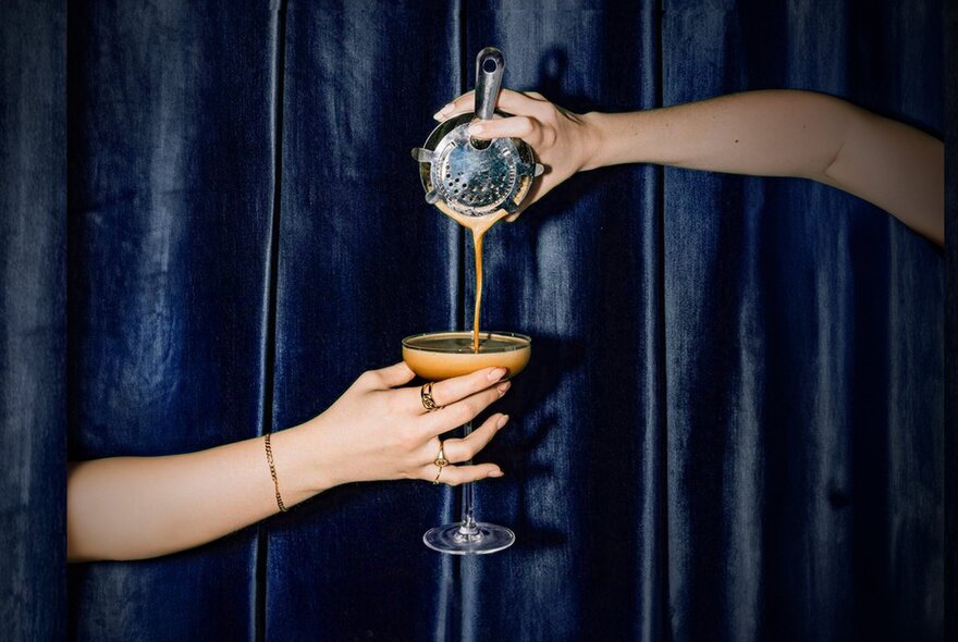 A hand pouring liquid from a cocktail shaker into a martini glass that is being held by another hand underneath it, against a blue velvet curtain.