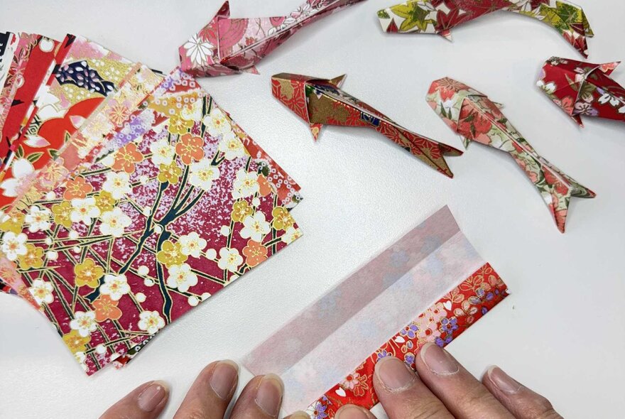 Small origami koi fish next to a pile of beautiful  Japanese floral origami paper.