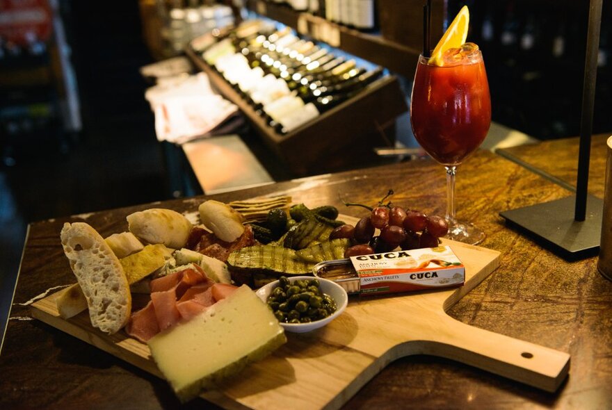 A charcuterie board with cheese, anchovies, olives and bread, next to a cocktail with a row of wine in the background. 
