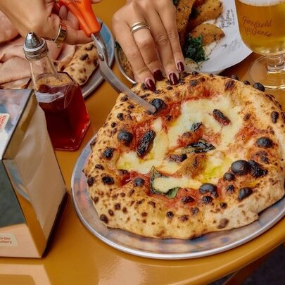 Someone cutting a margherita pizza with scissors next to chilli oil and wine. 