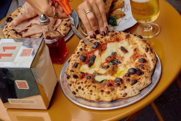 Someone cutting a margherita pizza with scissors next to chilli oil and wine. 
