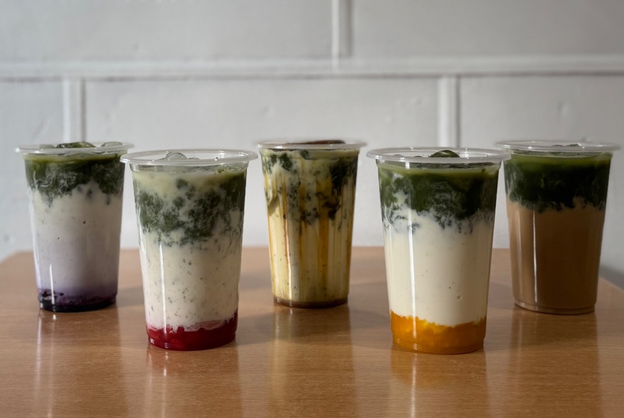 A line-up of five different matcha drinks in  clear plastic cups. 
