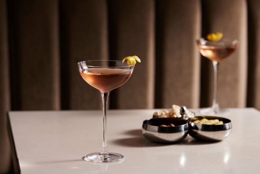 A table with two elegant cocktails and three small bowls of snacks. 