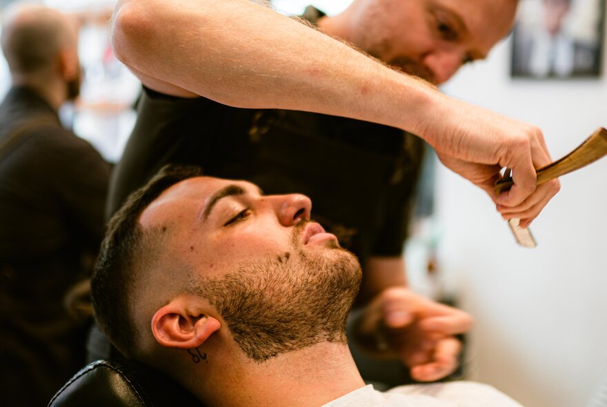A barber holds a blade over a client in a chair receiving a neck shave. 