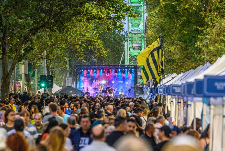 Crowds thronging a street with leafy trees, tent stalls and stage with lighting and live performance.
