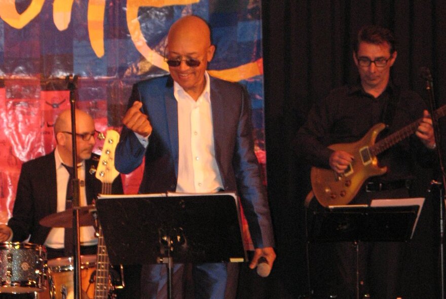 Bald jazz singer wearing sunglasses clicking fingers at a music stand, performing on stage with drummer and guitarist.