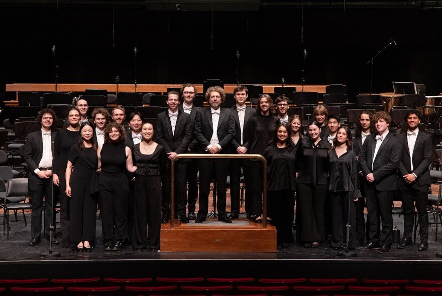 The ANAM Orchestra standing on stage without their instruments, dressed in black, behind conductor Asher Fisch on a wooden podium.