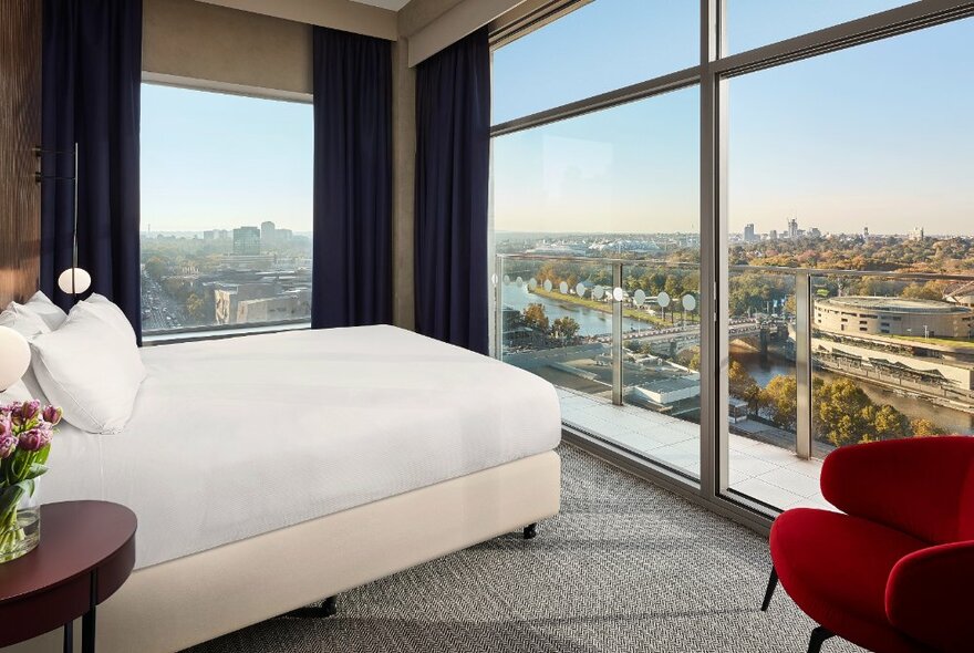 A hotel room with a white bed, and red armchair and large floor to ceiling windows overlooking the Yarra River and horizon.