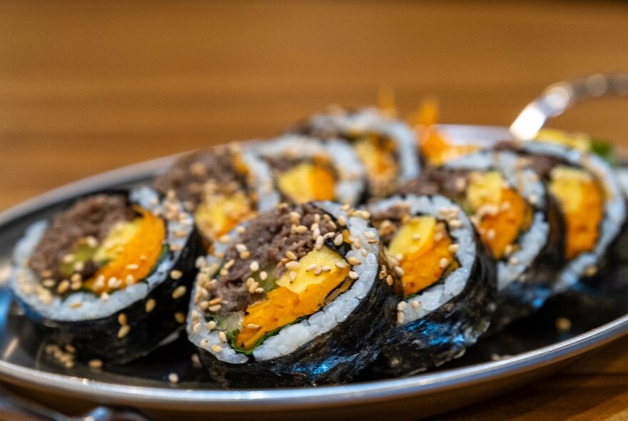 A silver tray of sliced gimbap wrapped in seaweed, filled with beef and sprinkled with sesame seeds.