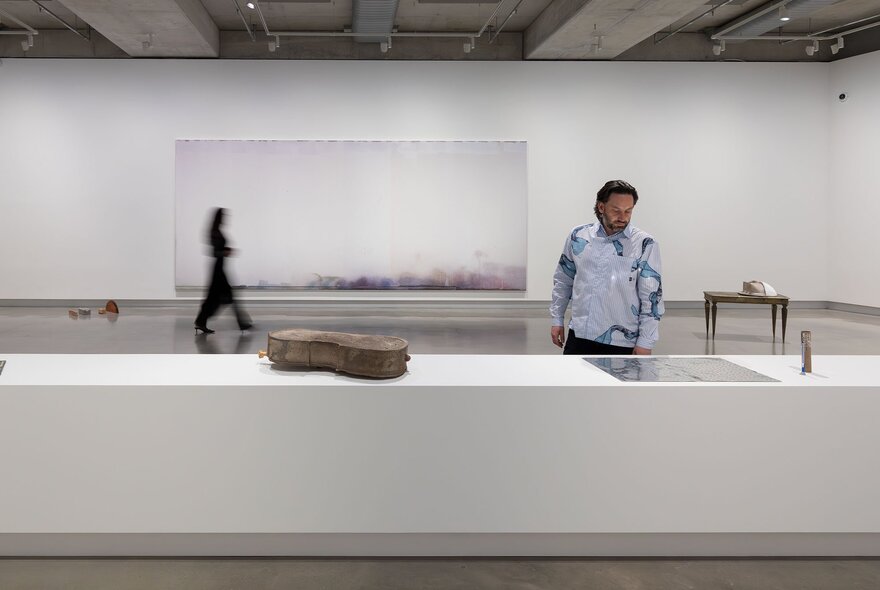 A large white room in a gallery, with a person looking at an object on a table, while in the background a person is walking past a large artwork on the wall.