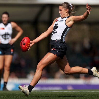 A Carlton AFLW player running on the field about to kick a red football that has just left her hands.