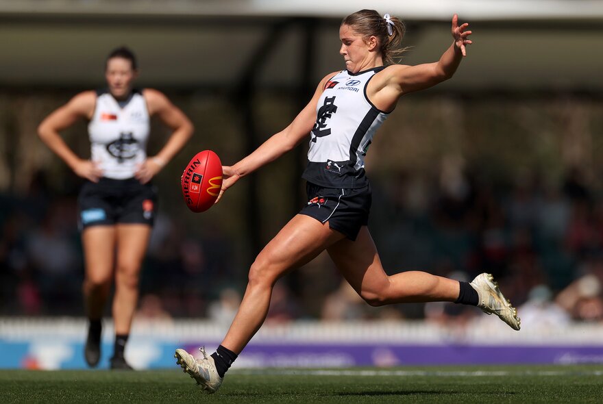 A Carlton AFLW player running on the field about to kick a red football that has just left her hands.