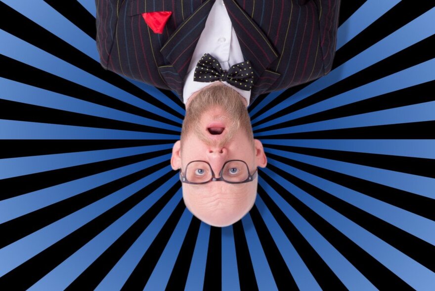 A man in a dinner suit and glasses upside down against a blue and black sun ray striped background. 