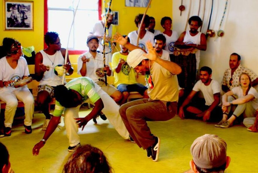 People participating in a Capoeira Angola class, with two people dancing and moving in the centre of a seated group of onlookers. 