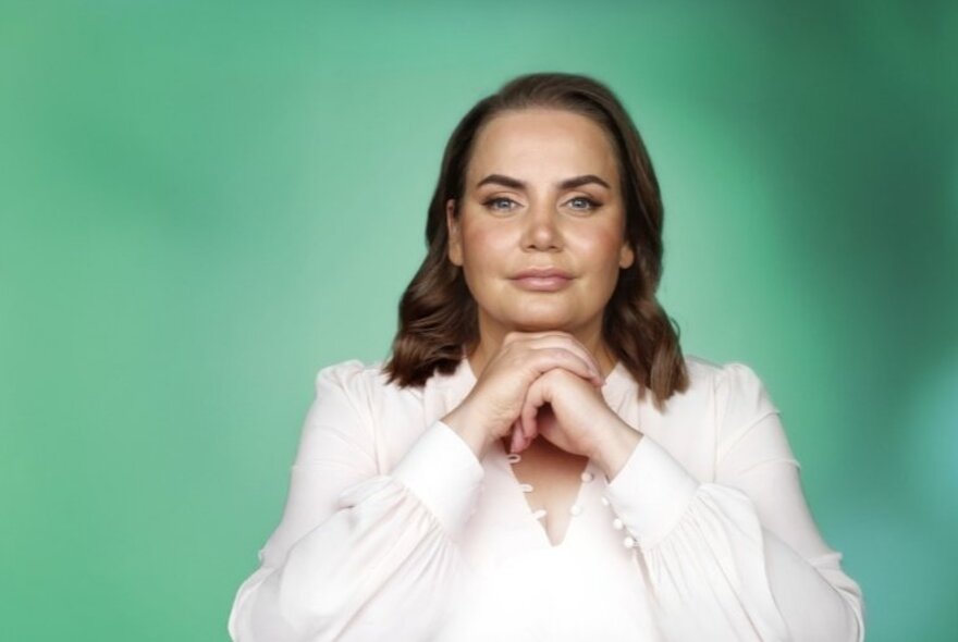 Tennis commentator and champion Jelena Dokic. sits smiling serenely with her hands clasped under her chin against a green background. 