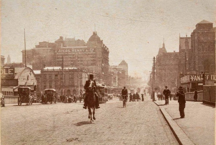 A sepia-toned vintage photograph of Swanston Street in Melbourne during the late 1800s with a person riding a horse down the wide road and Victorian-era buildings and horse-drawn carriages lining the street.