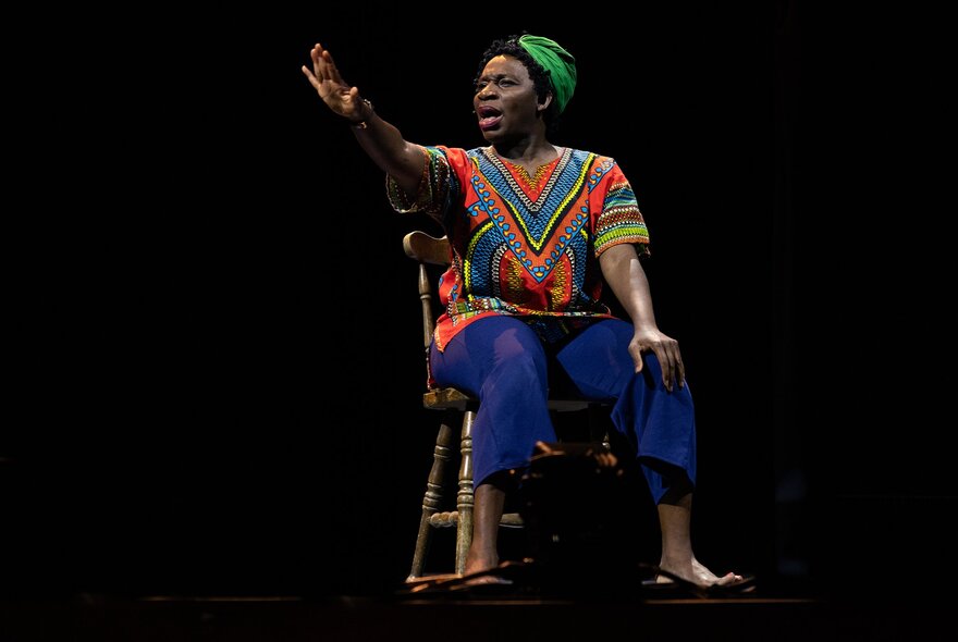 A woman on stage playing reknowned jazz singer and civil rights activist, Nina Simone, singing as she sits on a wooden chair.